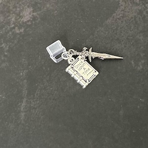 May include: A silver-colored charm featuring a miniature book with the words "Once upon a Time" and a small sword. The charm is attached to a clear plastic clasp. The charm is on a dark gray surface.