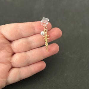 May include: A gold-colored earring featuring a small sword pendant, a faux pearl, and a clear clip. The sword has intricate details. The earring is held in a hand against a dark background.
