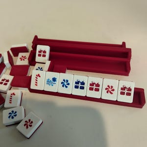 May include: A set of white mahjong tiles with red and blue engraved symbols, including floral and geometric patterns, arranged in and around two dark red plastic tile racks. The tiles feature various markings in red and blue ink.