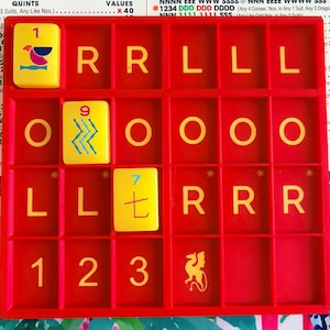 May include: A red plastic game rack with a grid of slots containing yellow tiles. The tiles feature various symbols, including the numbers 1, 9, and 7, letters R, L, and O, a bird icon, a dragon silhouette, and blue wavy lines.