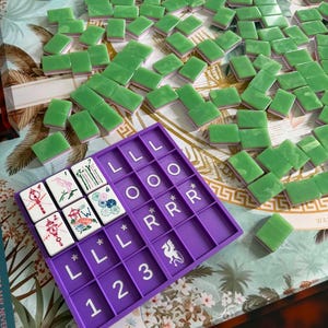 May include: A purple plastic Mah Jongg card holder with white letter and number labels, containing six white tiles with colorful illustrations. Scattered around the holder are numerous green and white tiles on a patterned surface with palm tree motifs.