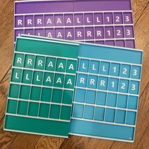 Könnte beinhalten: Drei farbenfrohe Wortspielbretter in Lila, Türkis und Blau werden gezeigt. Jedes Brett hat ein Raster aus Buchstaben- und Zahlenkacheln. Die Buchstaben buchstabieren Wörter wie "ALL" und "ARR". Die Bretter liegen auf einer Holzoberfläche.