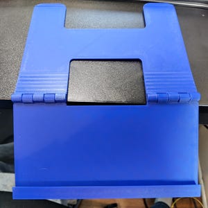 May include: A blue plastic book stand featuring a hinged design with a central rectangular cutout and a bottom ledge for support. The stand includes a black rectangular insert positioned within the central opening of the blue frame structure.