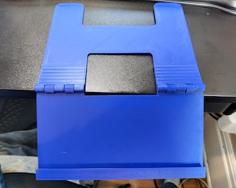 Peut inclure: Un support de livre en plastique bleu doté d'une conception articulée avec une découpe rectangulaire centrale et un rebord inférieur pour le soutien. Le support comprend un insert rectangulaire noir positionné dans l'ouverture centrale.