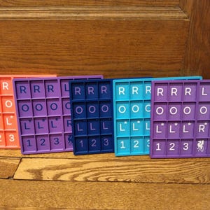 Op de afbeelding: Vijf kleurrijke, rechthoekige spelborden zijn naast elkaar geplaatst. Elk bord heeft een raster van letters en cijfers. De borden zijn in tinten oranje, paars, blauw, turquoise en violet. De tegels zijn wit.