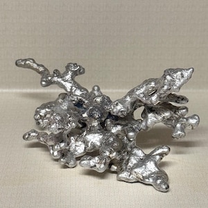 May include: A cast metal sculpture with a raw, organic, and irregular shape. The object features a shiny, silver-toned surface with a textured, bubbly, and porous appearance, resembling molten metal that has cooled into a complex, branching form.