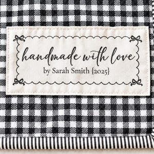 May include: A rectangular fabric label with a scalloped border and small bow accents at each corner, stitched onto a black and white gingham fabric. The label features the black printed text: handmade with love by Sarah Smith {2025}.