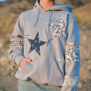 May include: Grey hoodie with star patches in various patterns. The hoodie has a front pocket and drawstring hood. Patches include floral, gingham, and lace designs. The model is wearing a denim skirt.