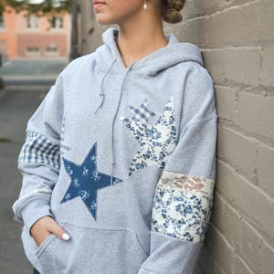 May include: A grey hoodie with star-shaped patches in blue floral and gingham patterns. The sleeves have fabric accents. The hoodie has a front pocket and a drawstring hood.