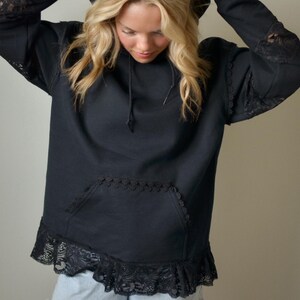 May include: Black hoodie with lace detailing on the sleeves, pocket, and hem. The hoodie has a drawstring hood and a front pocket. The sleeves have lace inserts. The hemline features a wide lace ruffle.