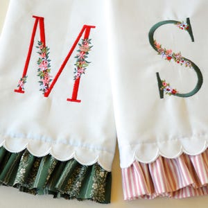 May include: Two white fabric towels featuring embroidered letters M and S. The letter M is red with floral accents, and the letter S is dark green with floral accents. Both towels have a scalloped bottom edge with ruffled fabric trim.