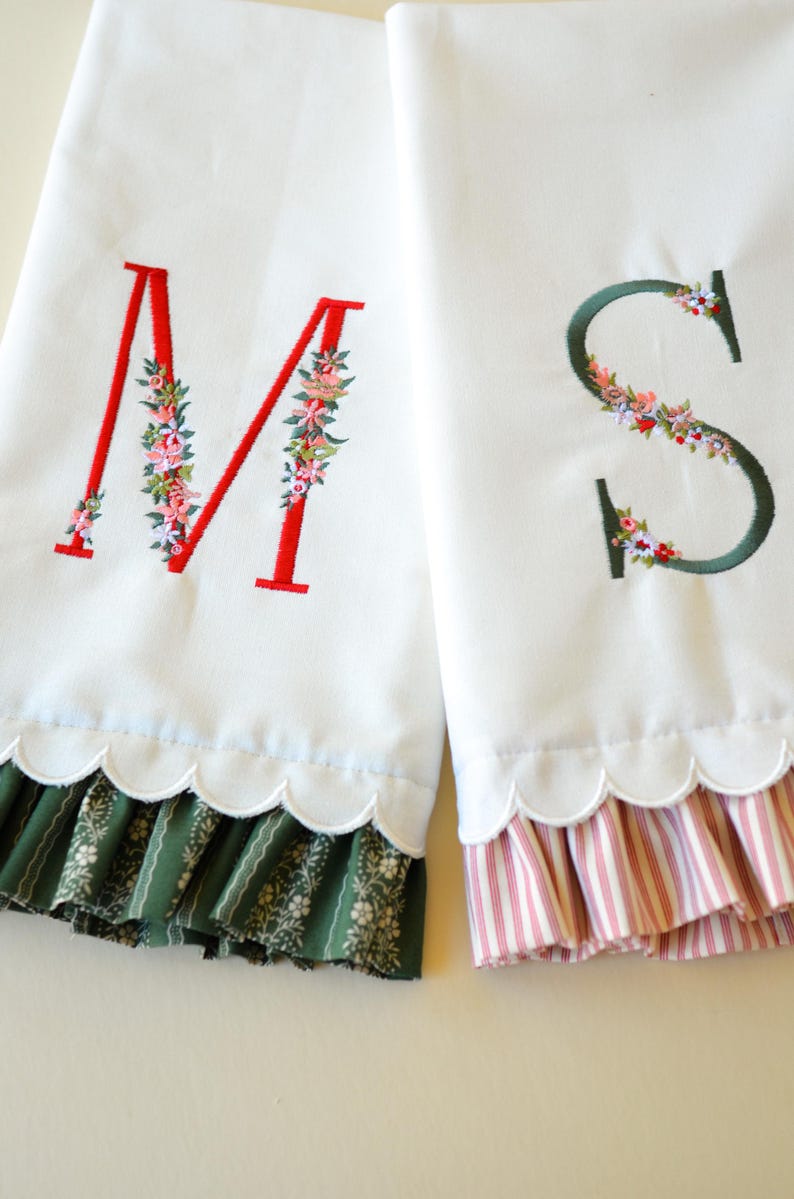 May include: Two white fabric towels featuring embroidered letters M and S. The letter M is red with floral accents, and the letter S is dark green with floral accents. Both towels have a scalloped bottom edge with ruffled fabric trim.
