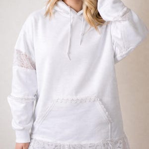 May include: White hoodie with lace accents on the sleeves, pocket, and hem. The hoodie has a drawstring and a front pocket. The lace detailing adds a delicate touch to the casual design. The garment is made of a soft, comfortable material.