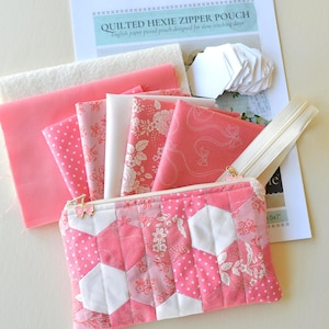 Puede incluir: Una bolsa con cremallera hexágono acolchada rosa con un estampado floral y de lunares, junto con trozos de tela, una cremallera y piezas de papel. El texto en el papel dice "QUILTED HEXIE ZIPPER POUCH".