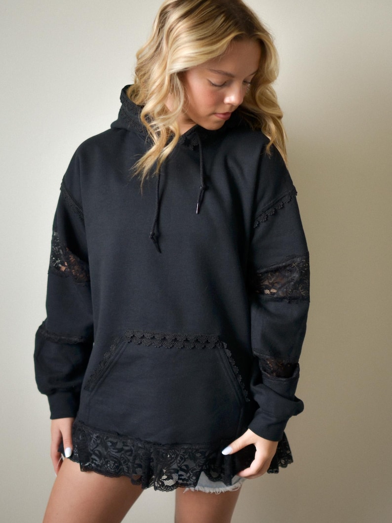 May include: Black hoodie with lace detailing on the sleeves, pocket, and hem. The hoodie has a drawstring hood and a front pocket. The lace adds a feminine touch to the casual design. A stylish and comfortable garment.