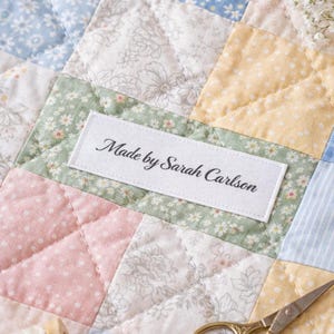 May include: A patchwork quilt with a variety of floral and patterned fabrics in pastel colors. The quilt features a label that reads "Made by Sarah Carlson". Gold scissors and a spool of thread are also visible.