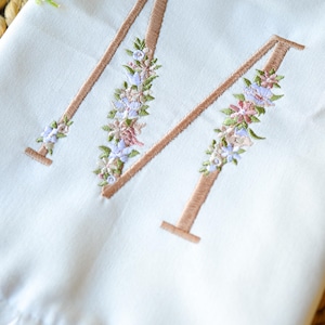 May include: A white fabric item featuring a large embroidered letter M in a tan thread color. The letter is decorated with a floral pattern consisting of small pink, blue, and green embroidered flowers and leaves along the vertical strokes.