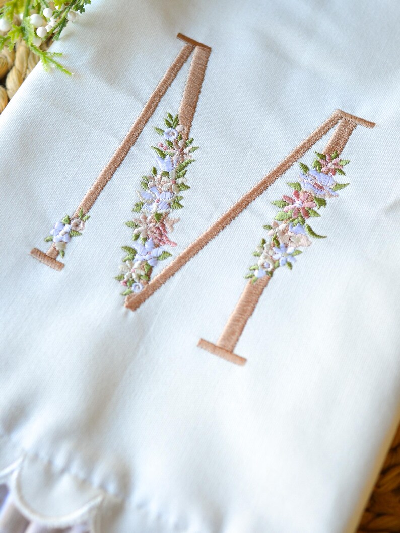 May include: A white fabric item featuring a large embroidered letter M in a tan thread color. The letter is decorated with a floral pattern consisting of small pink, blue, and green embroidered flowers and leaves along the vertical strokes.
