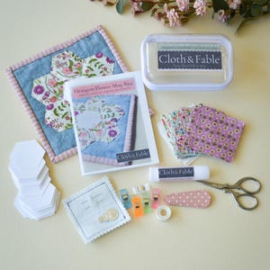 May include: A craft kit with a hexagon flower mug rug, paper piecing starter kit, and various sewing supplies. The kit includes a clear container, fabric pieces, scissors, clips, glue stick, and a needle holder. The brand name "Cloth & Fable" is visible.