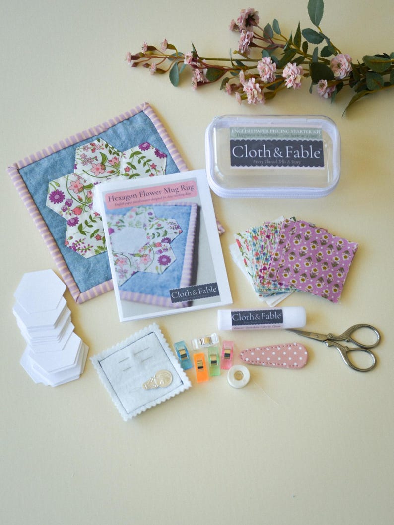 May include: A craft kit with a hexagon flower mug rug, paper piecing starter kit, and various sewing supplies. The kit includes a clear container, fabric pieces, scissors, clips, glue stick, and a needle holder. The brand name "Cloth & Fable" is visible.