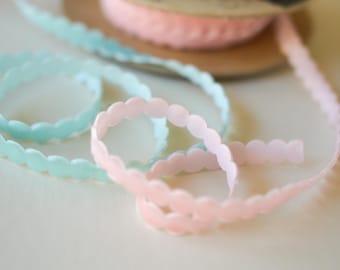 May include: A stack of spools with pastel-colored ribbons. The ribbons are light blue and pink with a scalloped edge. The spools are made of cardboard. The ribbons are unspooled and lying on a white surface.