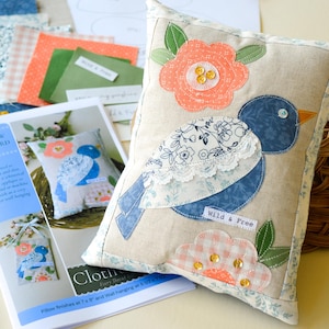 Peut inclure: Un coussin décoratif avec un appliqué d'oiseau bleu, une fleur pêche et un nuage rose à carreaux. Le coussin est beige avec une bordure florale et le texte "Wild & Free". Inclut un patron et des échantillons de tissu.