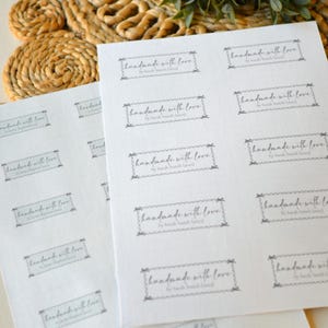 Op de afbeelding: Vellen witte etiketten bedrukt met de tekst "handmade with love" in een decoratief kader. De etiketten zijn gerangschikt op een licht oppervlak, met een geweven mat en groen op de achtergrond.