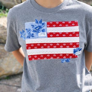 Puede incluir: Camiseta gris con un diseño de bandera americana de retazos. La bandera presenta rayas rojas y blancas con detalles de encaje floral, un campo floral azul y los números "1776" y "2026".