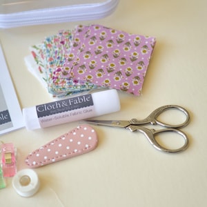 May include: A craft kit with fabric squares, scissors, glue, and a hair clip. The fabric squares have floral patterns in pink and other colors. The scissors are silver, and the glue stick is labeled "Water-Soluble Fabric Glue". The kit includes a clear zippered case.