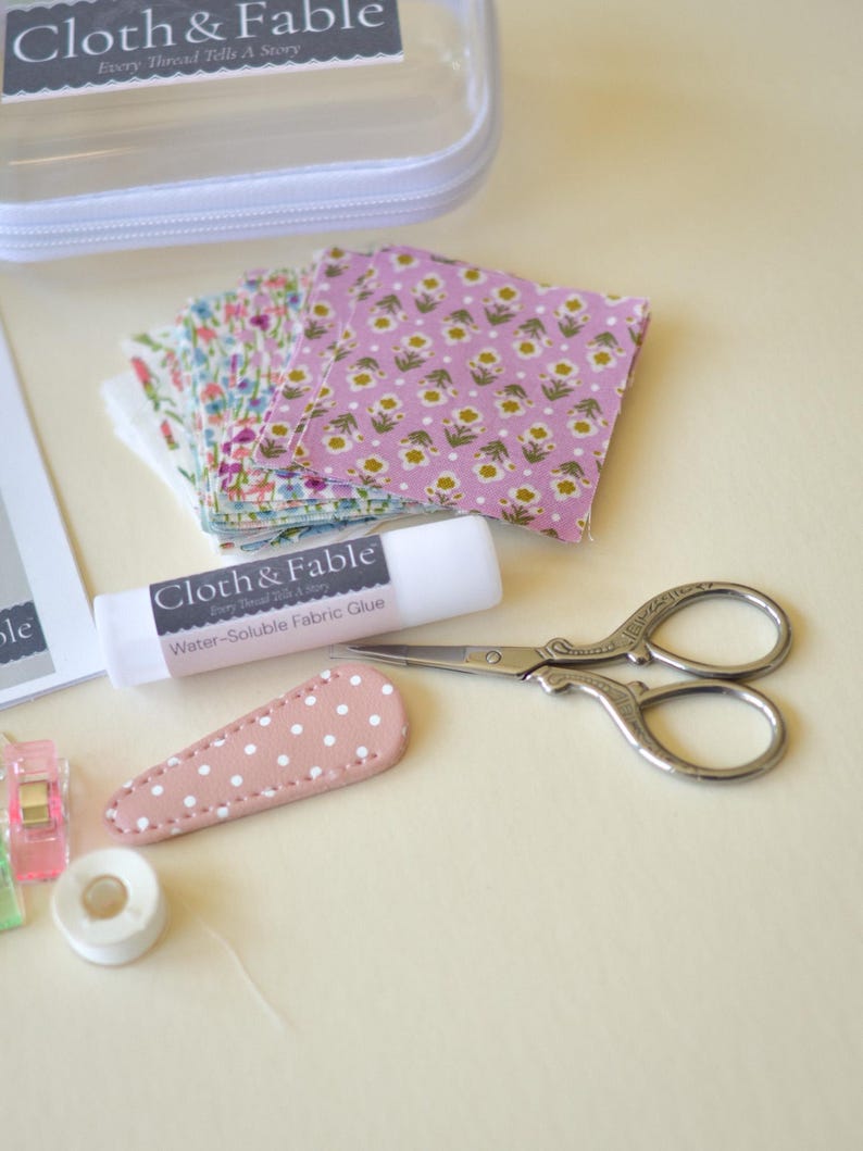 May include: A craft kit with fabric squares, scissors, glue, and a hair clip. The fabric squares have floral patterns in pink and other colors. The scissors are silver, and the glue stick is labeled "Water-Soluble Fabric Glue". The kit includes a clear zippered case.