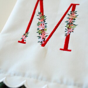 May include: A white fabric towel featuring a large, embroidered red letter M. The letter is decorated with intricate floral embroidery in shades of pink, green, and white. The bottom edge of the towel has a scalloped border design.