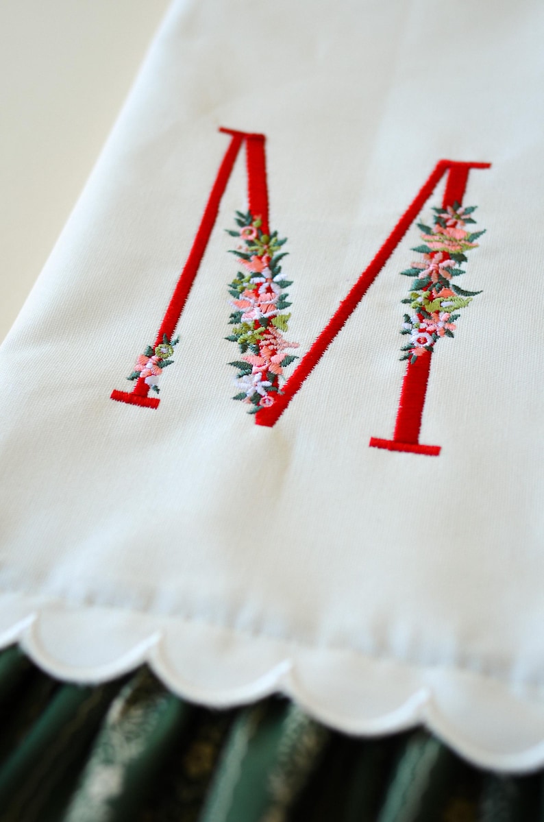 May include: A white fabric towel featuring a large, embroidered red letter M. The letter is decorated with intricate floral embroidery in shades of pink, green, and white. The bottom edge of the towel has a scalloped border design.