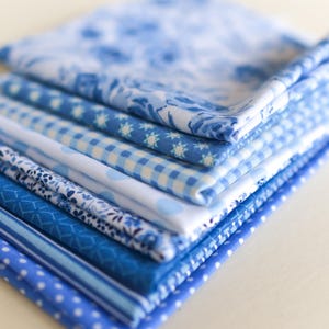 May include: A stack of various blue and white patterned fabrics. The fabrics include floral, gingham, polka dot, and striped designs. The fabrics are neatly folded and stacked, showcasing a range of shades from light to deep blue.