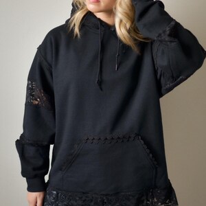 May include: Black hoodie with lace detailing on the sleeves, pocket, and hem. The hoodie has a drawstring hood and a front pocket. The lace adds a touch of elegance to the casual design. The hoodie is a comfortable and stylish garment.