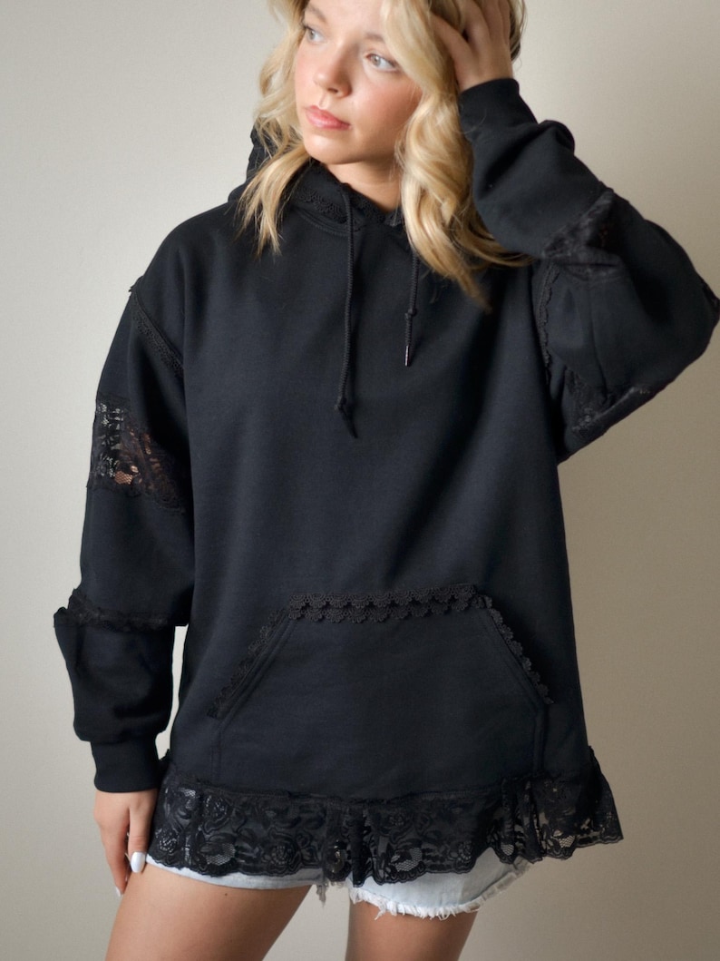 May include: Black hoodie with lace detailing on the sleeves, pocket, and hem. The hoodie has a drawstring hood and a front pocket. The lace adds a touch of elegance to the casual design. The hoodie is a comfortable and stylish garment.