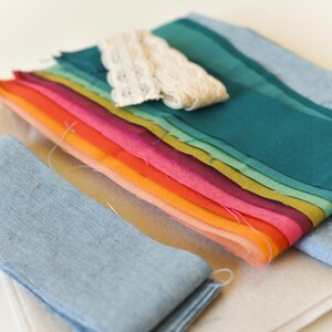May include: A collection of colorful fabric swatches in a rainbow of hues, including shades of teal, pink, orange, and yellow. A piece of cream-colored lace and two blue fabric strips are also present. The fabrics are arranged on a light beige surface.