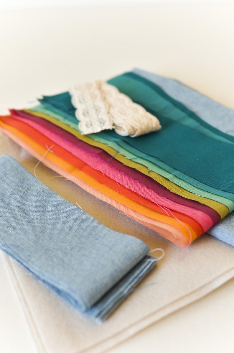 May include: A collection of colorful fabric swatches in a rainbow of hues, including shades of teal, pink, orange, and yellow. A piece of cream-colored lace and two blue fabric strips are also present. The fabrics are arranged on a light beige surface.