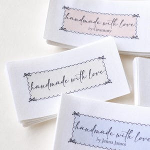 Op de afbeelding: Close-up van verschillende witte stoffen labels met de tekst "handmade with love" in cursief schrift. Elk label heeft een geschulpte rand en een gekleurde rechthoek met een naam. De labels zijn opgestapeld.