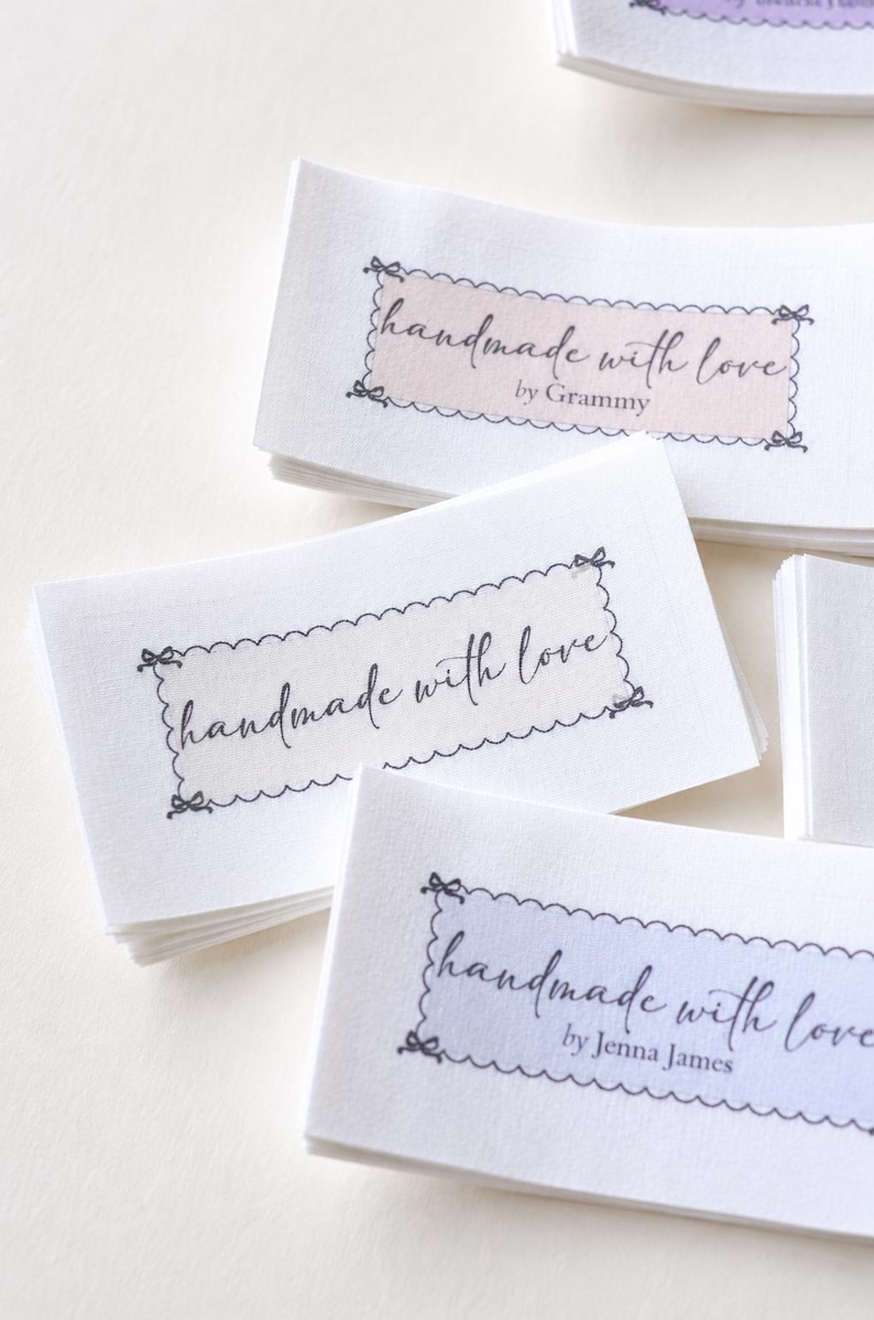 Op de afbeelding: Close-up van verschillende witte stoffen labels met de tekst "handmade with love" in cursief schrift. Elk label heeft een geschulpte rand en een gekleurde rechthoek met een naam. De labels zijn opgestapeld.