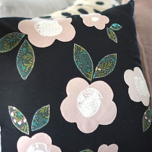 May include: Black decorative pillow with pink floral appliqués and green leaves. The flowers have white lace centers. The pillow is on a gray surface, with other pillows in the background.