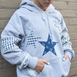 May include: A gray hooded sweatshirt with star appliqu&eacute;s. The stars are in blue floral and gingham patterns. The sleeves have gingham and floral patches. The sweatshirt has a front pocket and a drawstring hood.
