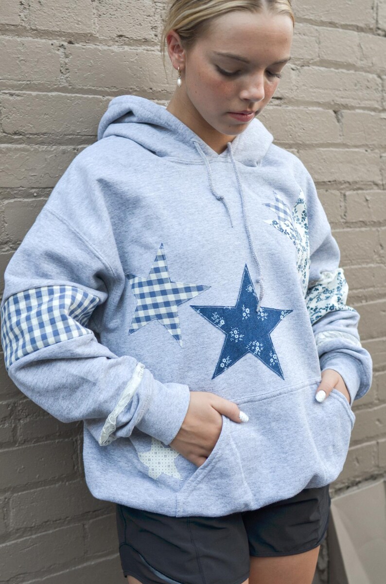 May include: A gray hooded sweatshirt with star appliqu&eacute;s. The stars are in blue floral and gingham patterns. The sleeves have gingham and floral patches. The sweatshirt has a front pocket and a drawstring hood.