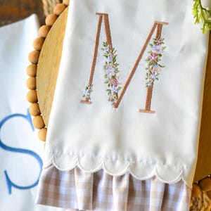 May include: A white kitchen towel featuring a large embroidered letter M in a copper-colored thread, decorated with small floral accents in pink, blue, and green. The bottom edge has a scalloped trim and a beige and white gingham fabric ruffle.