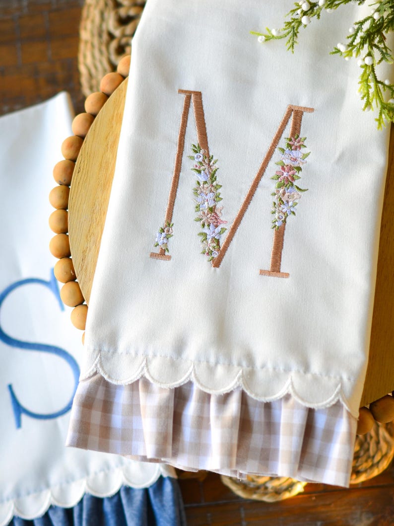 May include: A white kitchen towel featuring a large embroidered letter M in a copper-colored thread, decorated with small floral accents in pink, blue, and green. The bottom edge has a scalloped trim and a beige and white gingham fabric ruffle.