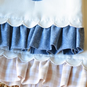 May include: A white fabric item featuring a blue embroidered letter S, finished with two scalloped edges. The top layer has a blue floral patterned ruffle, while the bottom layer features a light beige and white gingham checkered ruffle trim.