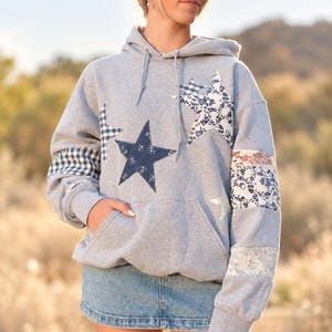 May include: A light gray hooded sweatshirt featuring a front kangaroo pocket and long sleeves. The garment is decorated with various star-shaped patches in blue and white floral, gingham, and denim patterns, along with white lace sleeve accents.