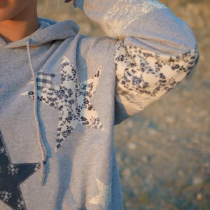 May include: Gray hoodie with star appliqu&eacute;s. The hoodie features a large star design on the front, with floral and gingham patterns. The sleeves have lace and floral fabric details. A drawstring hood and a front pocket complete the design.