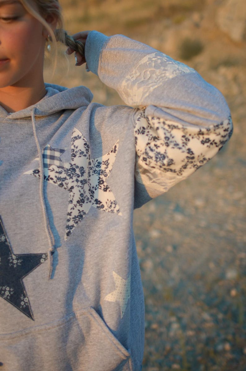 May include: Gray hoodie with star appliqu&eacute;s. The hoodie features a large star design on the front, with floral and gingham patterns. The sleeves have lace and floral fabric details. A drawstring hood and a front pocket complete the design.