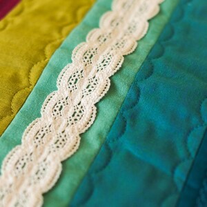 May include: Close-up of a colorful quilt with a white lace trim. The quilt features sections of teal, green, and magenta fabric. The lace is scalloped and adds a delicate touch to the handmade textile.