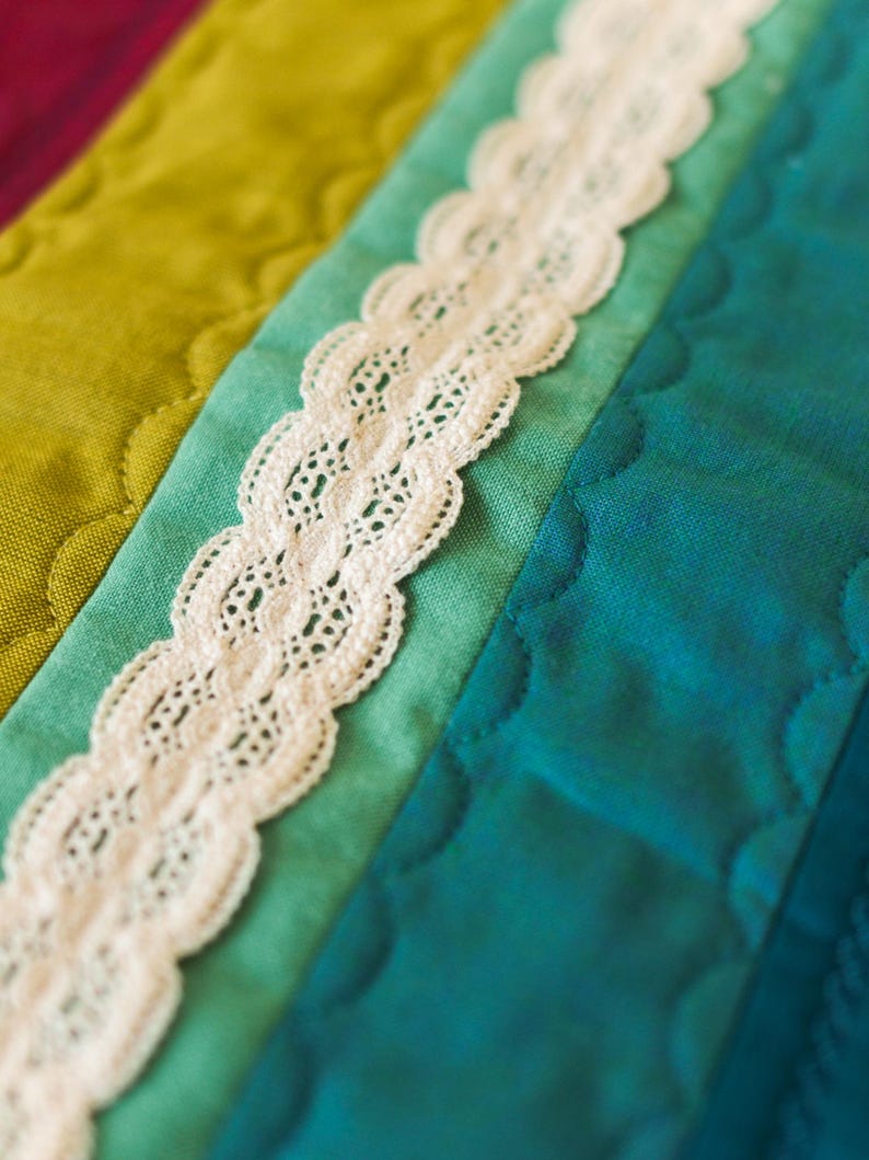 May include: Close-up of a colorful quilt with a white lace trim. The quilt features sections of teal, green, and magenta fabric. The lace is scalloped and adds a delicate touch to the handmade textile.